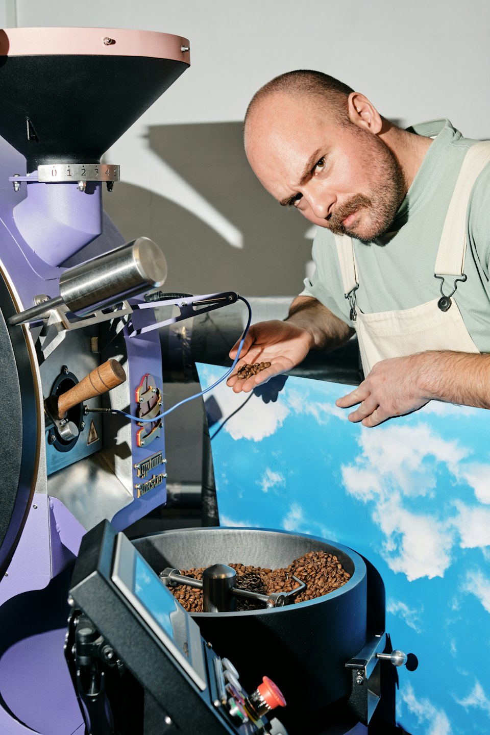 Ein bärtiger Mann inspiziert geröstete Kaffeebohnen neben einer Röstmaschine.